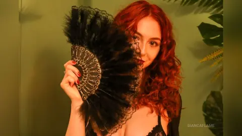 Redheaded woman in black lace outfit holding a large black lace fan in a moody, tropical setting.
