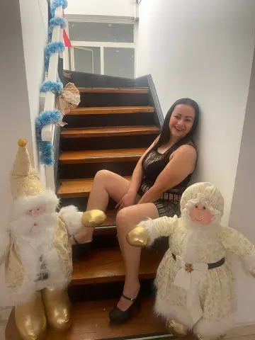 Smiling woman in a black dress sitting on wooden stairs next to large white and gold Santa decorations.