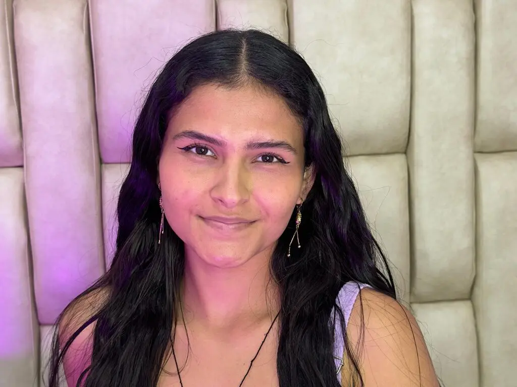 Smiling young woman with long dark hair wearing gold earrings in a cozy, soft-lit room.