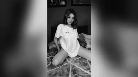 Young woman in a white oversized t-shirt sitting on a bed in a moody black and white indoor setting.