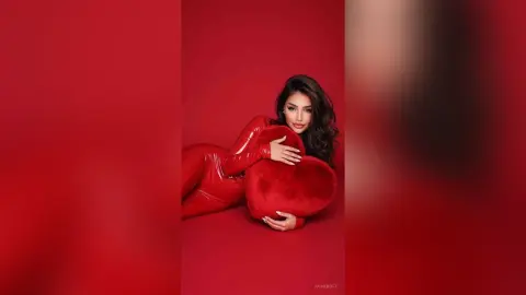 Glamorous woman in a red bodysuit reclining against a red heart pillow in a romantic red studio setting.
