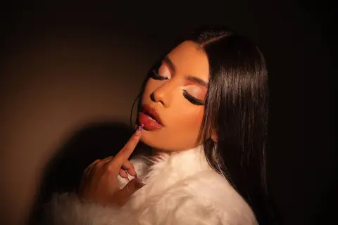 Glamorous woman with long dark hair and dramatic eye makeup wearing a white faux fur coat in moody lighting.