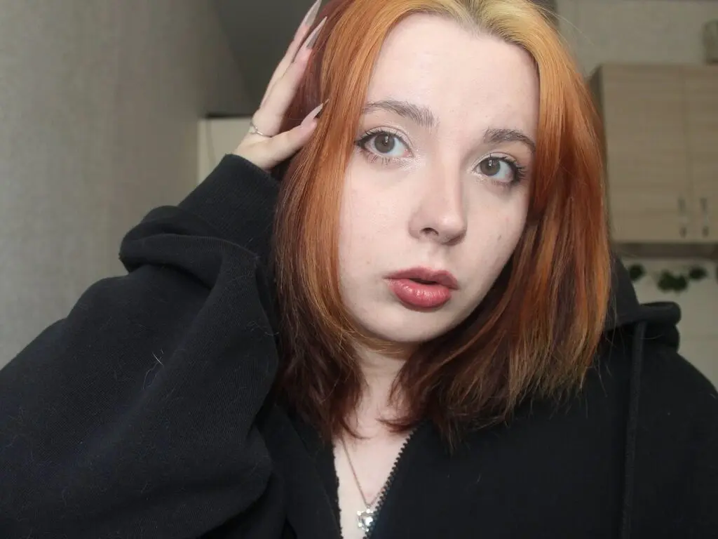 Redheaded woman in a black hoodie looking thoughtfully at the camera in a cozy indoor setting.