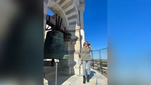 Woman in a beige coat and sunglasses standing by a large historic bell under a clear blue sky.