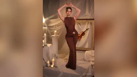 Elegant woman in a brown sleeveless gown posing gracefully in a warm, candlelit room with a vintage violin.