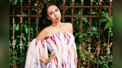 Elegant woman in a white floral off-the-shoulder dress posing by a lush green garden fence.