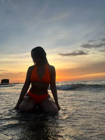 Silhouette of a woman in an orange bikini sitting in ocean waves during a golden sunset at the beach.
