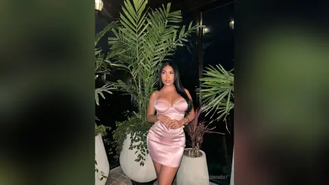 Glamorous woman in a pink satin mini dress posing by tropical plants in a moody evening setting.