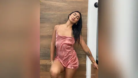 Elegant woman in a pink satin slip dress leaning against a wooden wall in a serene, moody setting.