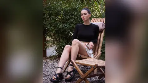 Young woman in a black long-sleeve top and black sandals relaxing on a wooden lounge chair in a lush garden.