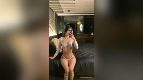 Young woman in a white long-sleeve crop top and bikini bottoms posing in a modern bedroom setting.