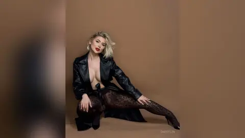 Glamorous blonde woman in a black leather jacket and leopard print tights posing against a brown studio backdrop.