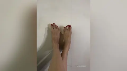 Close-up of bare feet with red nail polish resting against a plain white wall in a minimalist setting.