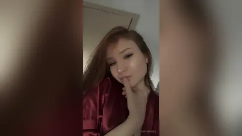 Young woman in a red satin pajama top posing with a thoughtful expression in a dimly lit room.
