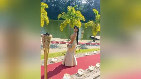 Elegant woman in a gold evening gown posing on a red carpet by a tropical lake with palm trees.