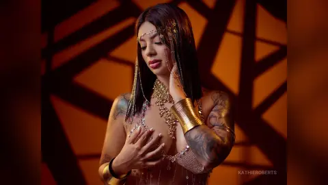 Glamorous woman in gold bohemian jewelry and sequined outfit posing against a warm, dramatic geometric background.