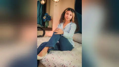 Smiling woman in a blue striped shirt and jeans sitting on a bed in a cozy, sunlit bedroom.