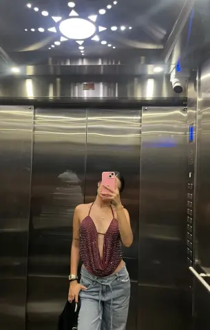 Young woman in a sparkly red halter top and light wash jeans taking a mirror selfie in a modern elevator.