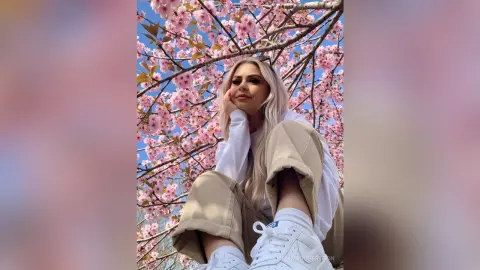 Blonde woman in white sweater and khaki pants relaxing under blooming pink cherry blossom trees in spring.