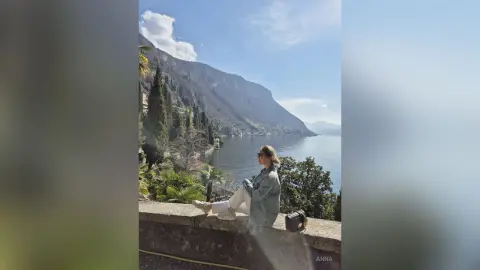 Woman in a light blue sweater sitting on a stone wall overlooking a scenic lake and mountains.