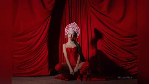 Elegant woman in a red corset and ornate white headpiece posing against dramatic red velvet curtains.