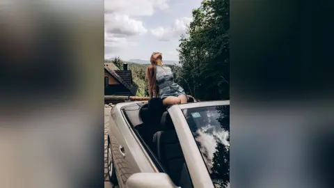 AdelinaKael sits in a convertible car, wearing a short blue dress and looking up toward the sky against a backdrop of trees and clouds.