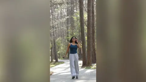 Smiling woman in a blue tank top and grey trousers walking through a sunny, lush pine forest.