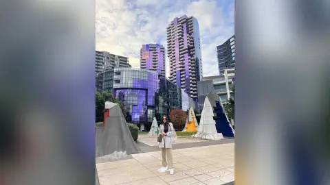 Stylish woman in a white blazer and tan trousers standing in a modern urban plaza with tall glass skyscrapers.
