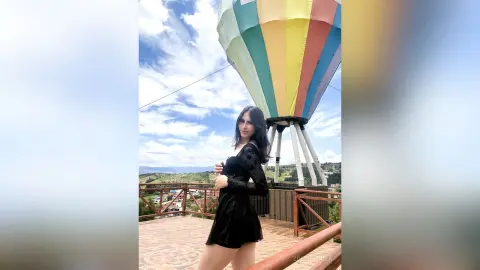 Stylish woman in a black velvet dress standing on a balcony with a colorful hot air balloon in the sky.