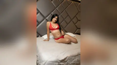 LauraMarcuzzi poses on a white bed wearing a red lace bra and matching red panties, leaning back against a grey padded headboard.