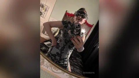 Smiling woman in a black lace dress and masquerade mask taking a mirror selfie in a cozy bedroom.