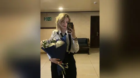 Blonde woman in a white shirt holding a large bouquet of flowers in a dimly lit room.