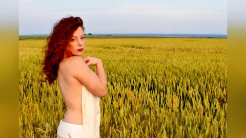 Redheaded woman in a white dress standing in a golden wheat field under a clear sky at sunset.