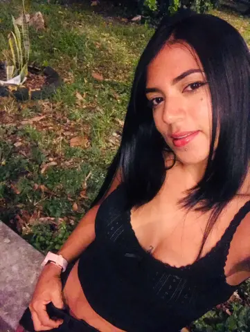 SusanaGlace with long black hair wearing a black lace camisole, posing outdoors in a grassy area.