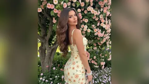 Glamorous woman in a floral sundress posing by a blooming pink rose bush in a sunny garden.