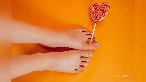 Close-up of bare feet resting on a bright orange background next to a red heart-shaped swirl lollipop.