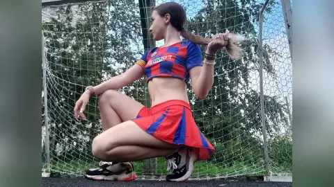 Young woman in a red and blue soccer uniform squatting outdoors near a sports net on a sunny day.