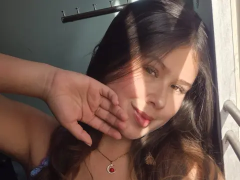 Young woman with long brown hair in sunlight wearing a delicate gold necklace in a bright, warm setting.