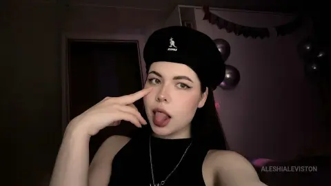 Young woman in a black sleeveless top and beanie making a playful expression in a dimly lit room.