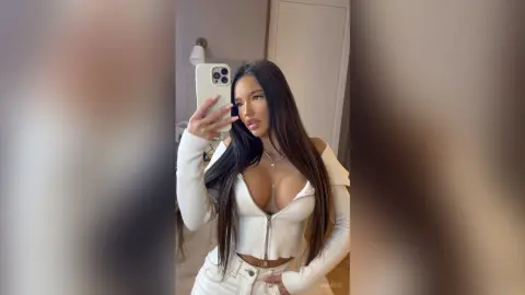 Young woman in a white long-sleeve crop top taking a confident mirror selfie in a bright room.
