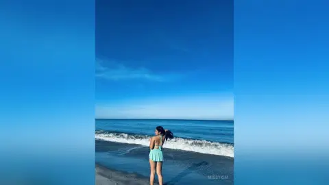 Young woman in a mint green bikini standing on a sunny beach facing the blue ocean waves.