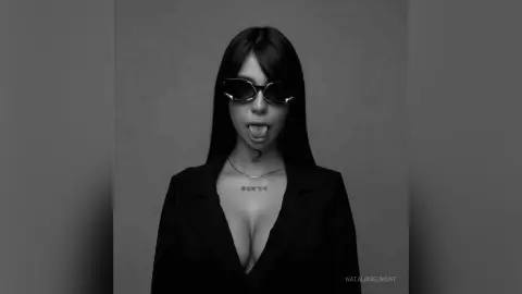 Edgy portrait of a woman in a black blazer and sunglasses sticking her tongue out against a gray background.