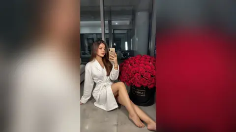 Young woman in a white silk robe sitting next to a large bouquet of red roses in a modern bright room.