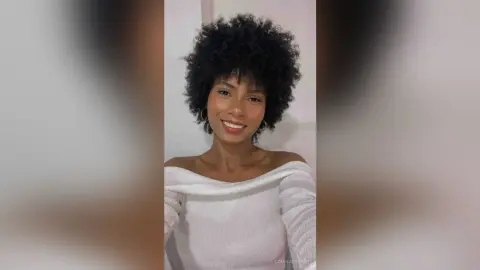 Smiling woman with curly afro hair wearing a white off-the-shoulder top in a bright indoor setting.