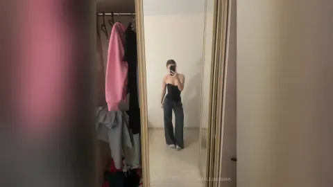 Young woman in a black tube top and grey wide-leg trousers taking a mirror selfie in a minimalist bedroom.