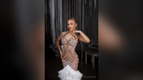 StellaFletcher poses in a sheer, beaded white bodysuit with a feathered skirt against a white string backdrop.