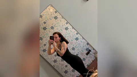 Woman in a black sleeveless dress taking a mirror selfie against a floral patterned wall in a modern room.