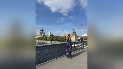 Joyful woman in a long purple dress raising arms on a bridge overlooking a historic cathedral under a blue sky.