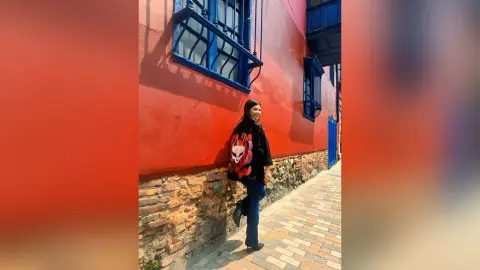Stylish woman in black outfit and backpack walking along a vibrant red building in a sunny urban alleyway.