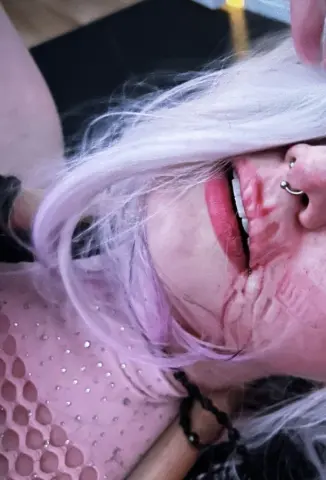 Woman with pastel lavender hair and pink makeup wearing a sparkly mesh top in a moody, close-up portrait.